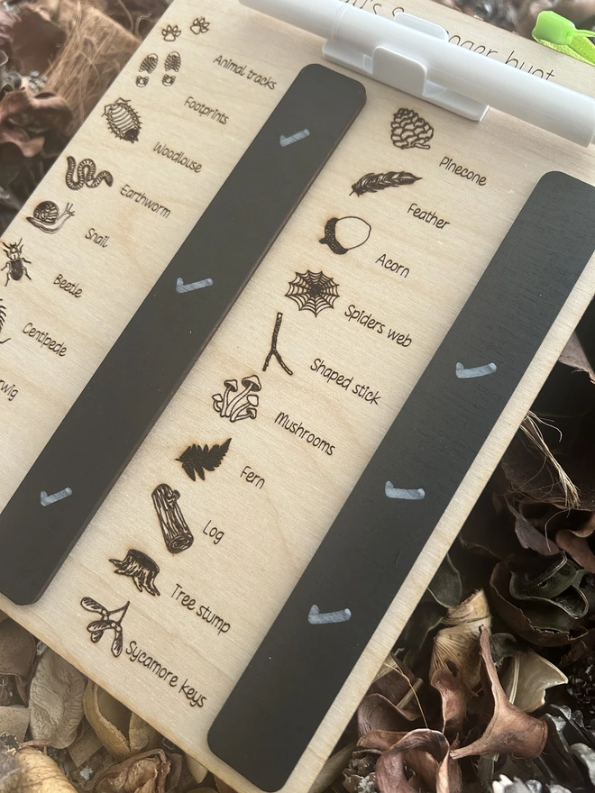 A personalised wooden scavenger hunt board titled “Holly’s Scavenger Hunt,” displayed on a bed of dried leaves and pinecones. The board features engraved illustrations and labels for natural items and minibeasts to find, including pinecone, feather, acorn, spider’s web, shaped stick, mushrooms, fern, log, tree stump, sycamore keys, animal tracks, footprints, woodlouse, earthworm, snail, beetle, centipede, earwig, butterfly, and ant. Two black wipe‑clean sections sit on the board, with a white marker attached by a green cord decorated with a small ladybird bead.