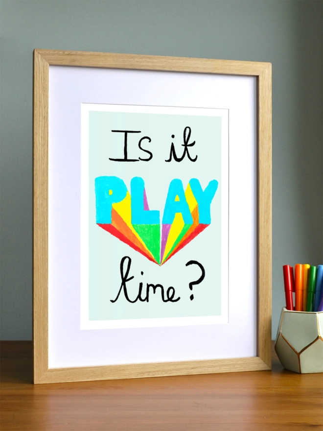 Art print saying 'Is it play time?' in a brown frame on a child's desk
