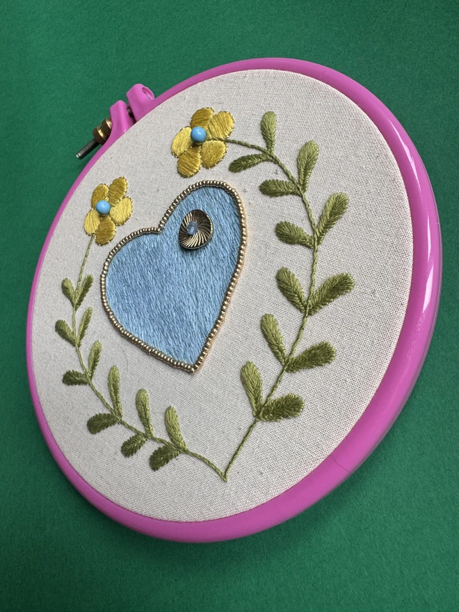 A pink embroidery hoop containing a blue stitched heart edged with gold beads surrounded by two yellow flowers on long stems