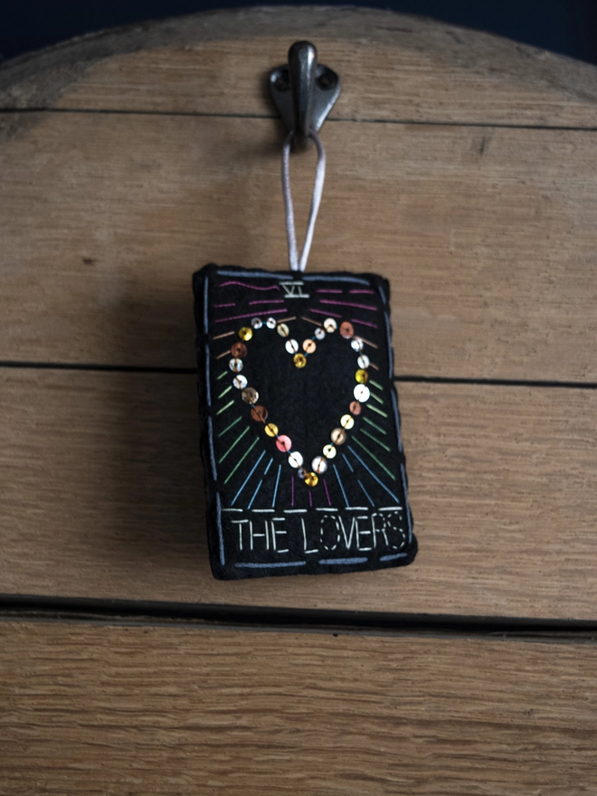 A handcrafted felt ornament with sequins and embroidery depicting "The Lovers" tarot card. Colourful embroidery with a heart shape. 