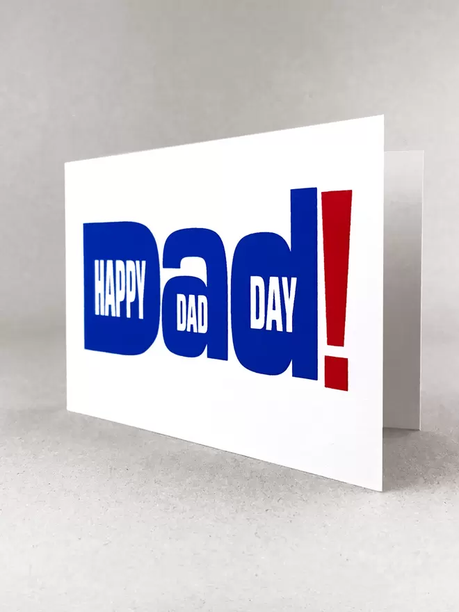 Studio shot of white landscape greetings card on light grey background, at a slight angle, so slightly open. The design is typography of the word Dad! With happy Dad Day written within the letters. The writing is printed in royal blue with an oversized exclamation mark in red to the right. 