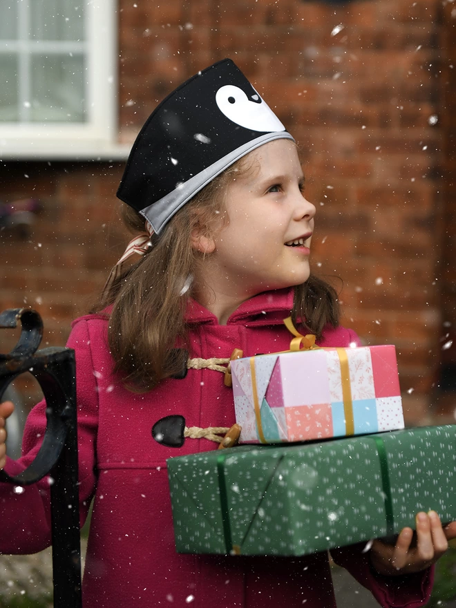 A young girl wearing a pink winter coat and a handmade penguin fabric crown stands outside in the snow while holding wrapped gifts.