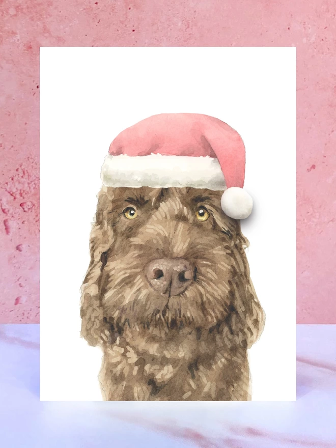 A Christmas card featuring a hand painted design of a chocolate cockapoo, stood upright on a marble surface surrounded by pompoms. 