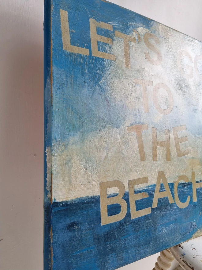 Upcycled art panel in blues and creams featuring a seascape and an image of a retro pin-up lady on a boat and the text reads ‘let’s go to the beach’ in a distressed, shabby chic style