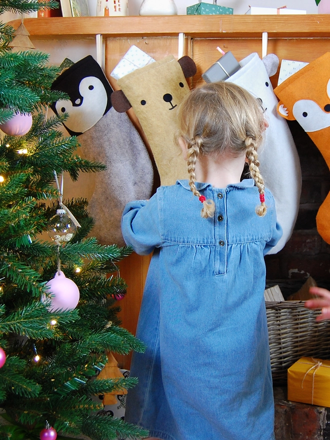 A young girl wearing a blue denim dress is standing in front of several animal stockings hanging on a wooden mantlepiece.