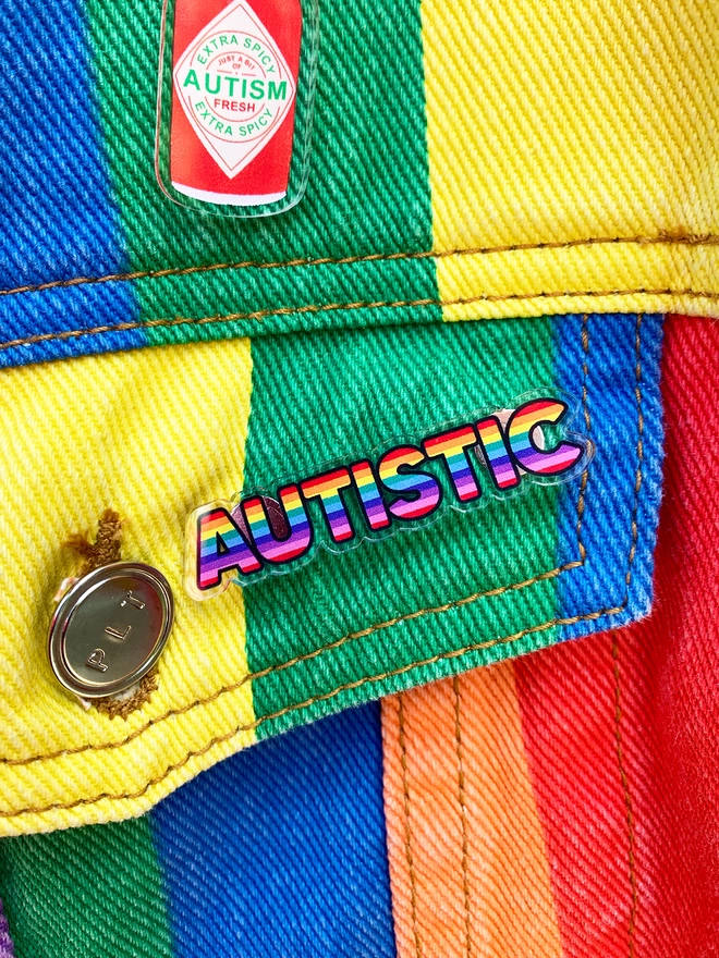 image shows an acrylic badge in the shape of the word 'autistic'. the letters are filled with horizontal rainbow stripes. the badge is pinned to the pocket of a rainbow striped denim jacket.