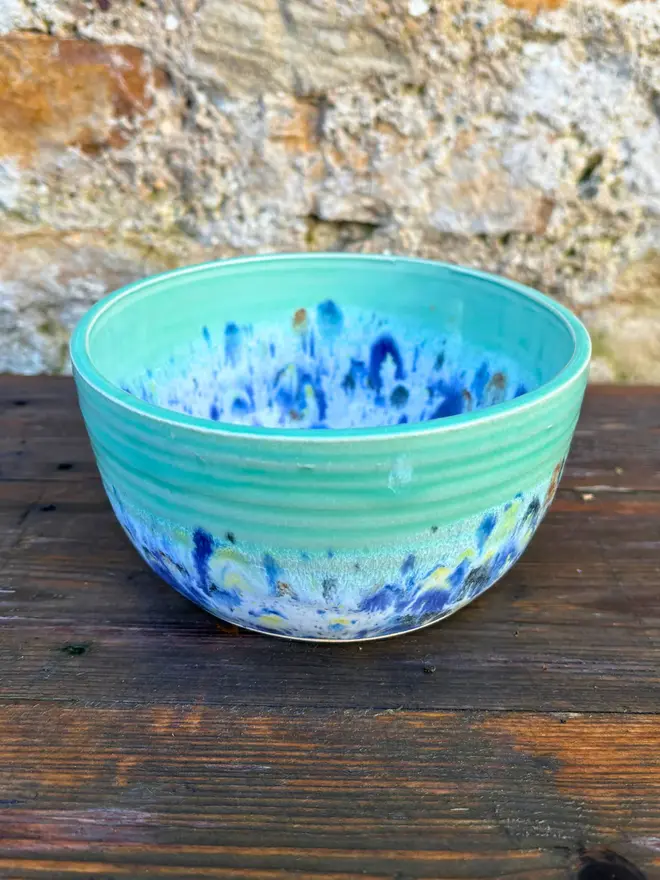 Shoreline Small Bowl, a colourful bowl placed on a wooden surface in front of a brick wall. 