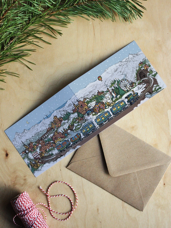 fold out greeting card with christmas town illustration flat on wooden table with foliage and string