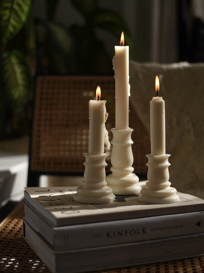 Baluster Christmas Candles (Set Of 2). Three glowing candlestick candles with soft wax drips on a stack of books, beside a cane chair and leafy houseplants. The warm candlelight creates a cosy, romantic mood for slow evenings at home.