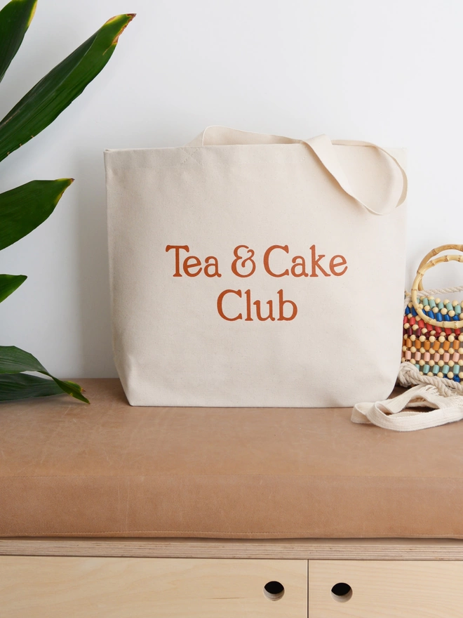 a large natural canvas tote bag bearing the words tea and cake club stood on a bench