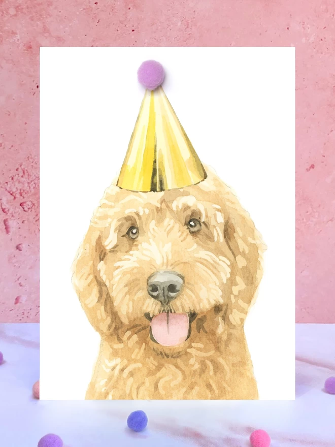 A greeting card featuring a hand painted design of a ruby cockapoo, stood upright on a marble surface surrounded by pompoms. 