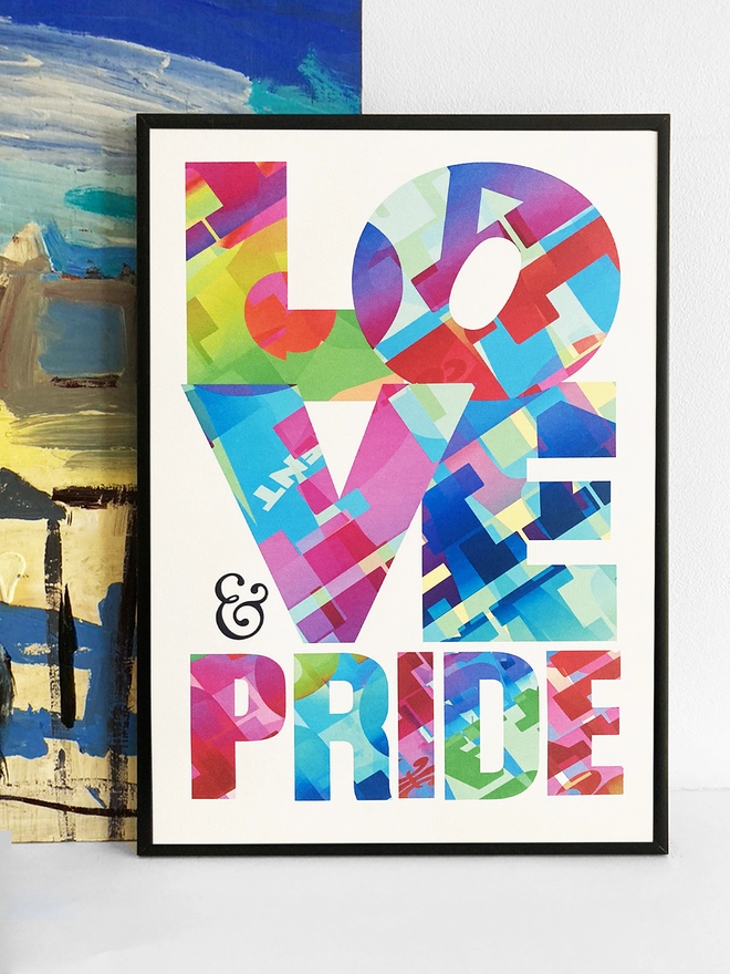 Framed multicoloured typographic print of “Love and Pride”  The print rests against a blue and yellow abstract painting.