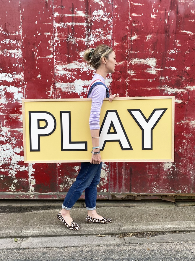 A person holding a yellow wooden sign which reads PLAY with one hand whilst walking in front of a red wall