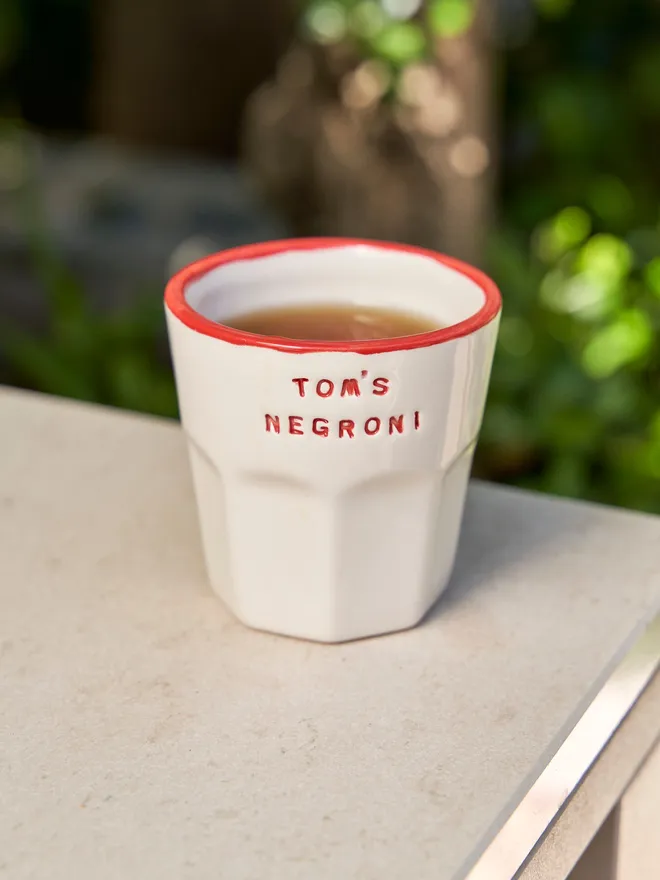 'Tom's Negroni' in a red font stamped on a ceramic cup with a red rim, on a table outside