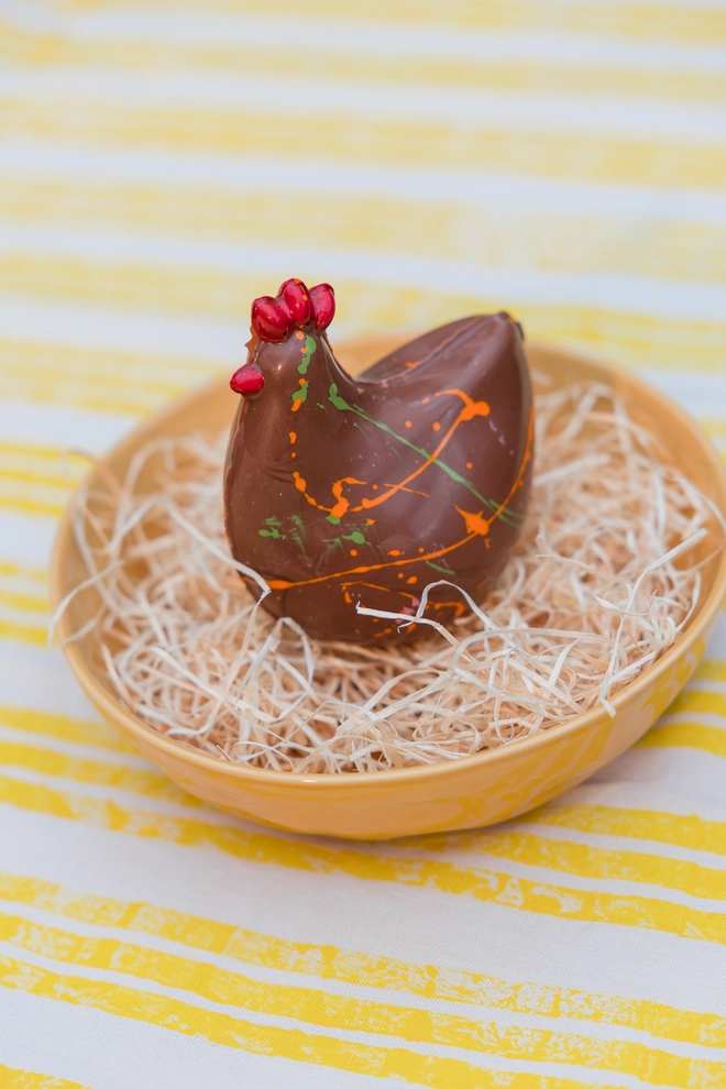 A milk chocolate Easter chicken from Nenette Chocolates sitting in  straw bedding in a yellow bowl, on a striped yellow and white tablecloth