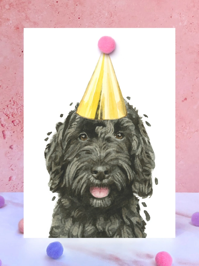 A greeting card featuring a hand painted design of a black cockapoo, stood upright on a marble surface surrounded by pompoms. 