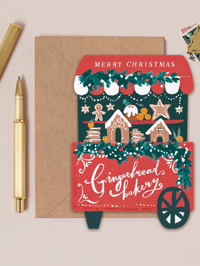 Red Gingerbread Cart Christmas card shaped like a gingerbread bakery market cart under a 'Merry Christmas' canopy, on a kraft envelope beside a gold pen