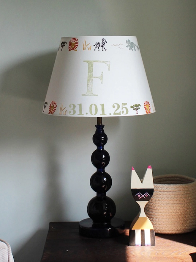 Safari lampshade with letter F, styled on side table in bright boys bedroom. Handmade safari lampshade for kids bedroom, personalised with letter F. Colourful empire light shade with safari animals, a green lampshade option for boys lampshade or kids bedroom design. The lamp is on a dark blue lamp base next to a cream arm chair with blue cushion and sheep skin. 