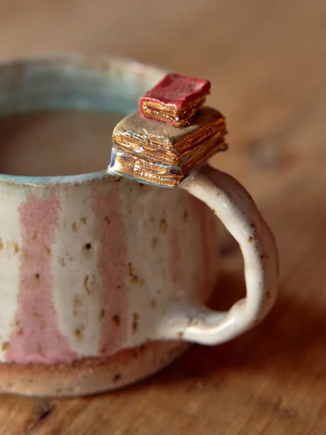 Close up of pink and white striped ceramic mug with book detail on the handle, pictured on a wooden table - emphasis on the book detail