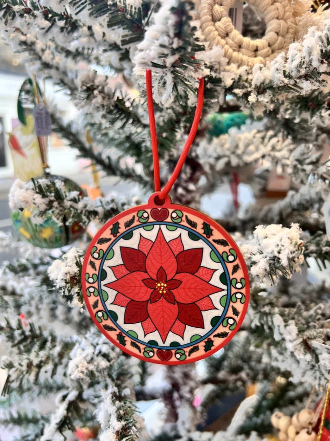 Multi coloured wooden tree decoration with a red poinsettia pattern at the centre. It is hanging by a red ribbon on a fake white christmas tree with other decorations. 