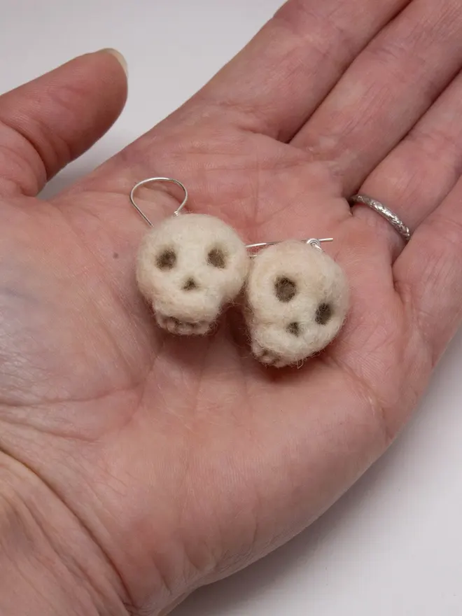 Two needle-felted skull earrings in the palm of a hand. Made by the artist Laura Cronin aka Bumble & Earwig