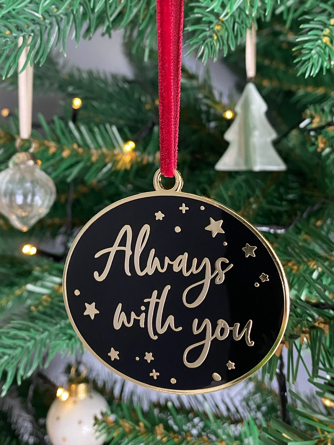 A black and gold enamel Christmas decoration, with the words “Always With You” surrounded by gold stars, hangs on a Christmas tree.
