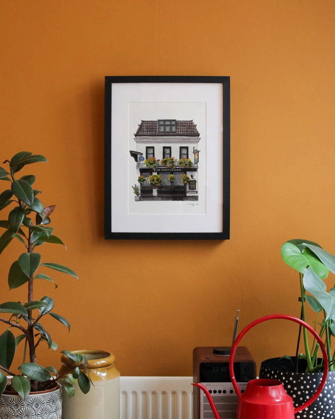 An illustration of "The Mayflower" pub in a black frame, centered on a warm orange wall above indoor plants, a vintage radio, and a red watering can.