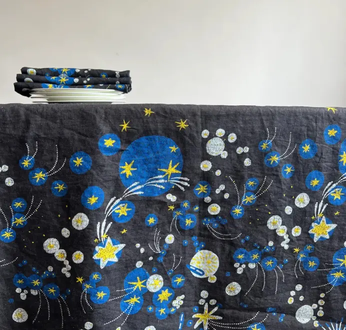Solaris Tablecloth In White Or Blue. Blue tablecloth with blue and yellow starburst motifs, styled with white plates and a matching napkin.