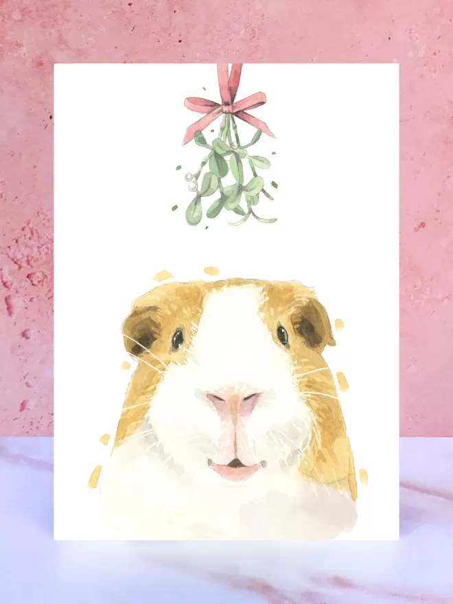 A Christmas card featuring a hand painted design of a Guinea Pig, stood upright on a marble surface.