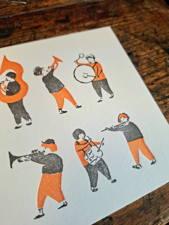 The cover of a greetings card lying open on a wooden surface, with an illustration of small orange and black figures playing musical instruments printed on the front