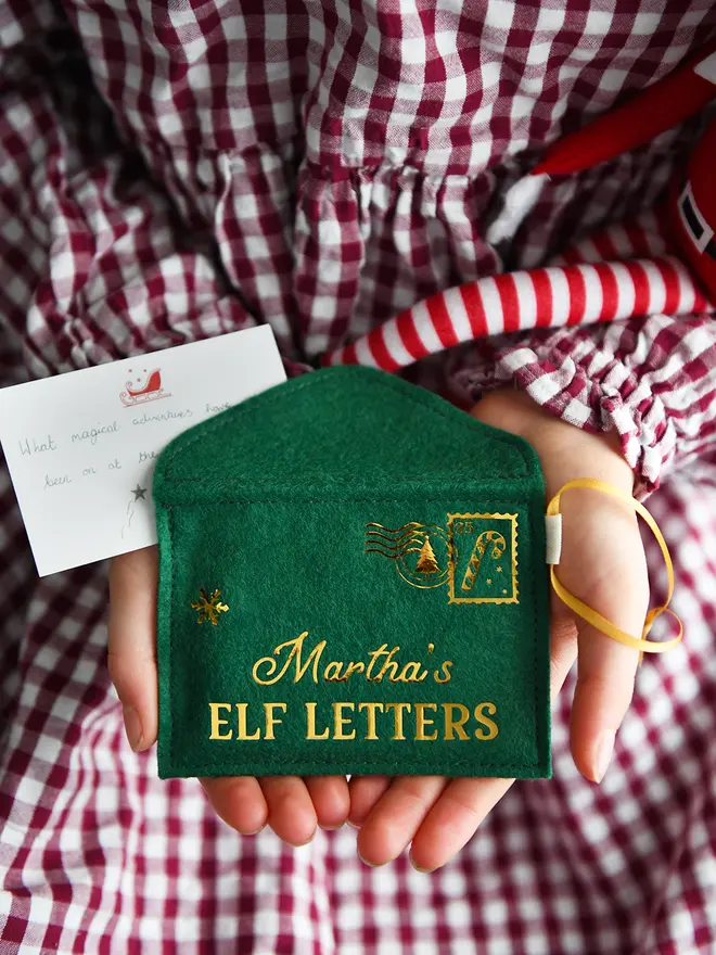 A child wearing a red gingham dress is holding a handmade green felt envelope with golden words reading "Martha's elf letters" and a candy cane design stamp in the corner. 