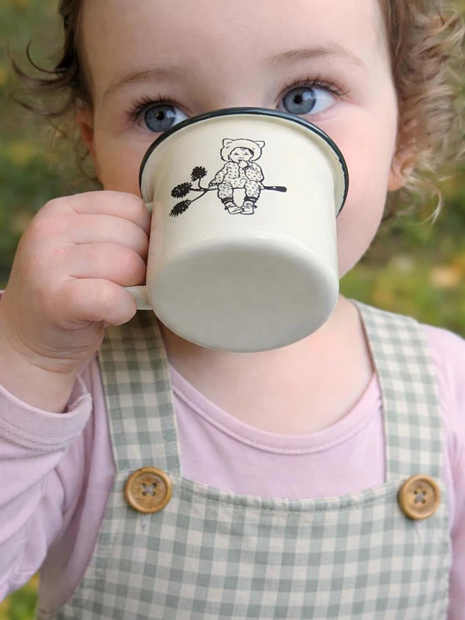 a sweet child drinking from a miniature enamel mug featuring a story book style illustration of a sweet pixie child sat on a pine cone branch cream in colour with a bottle green rim vintage retro in style