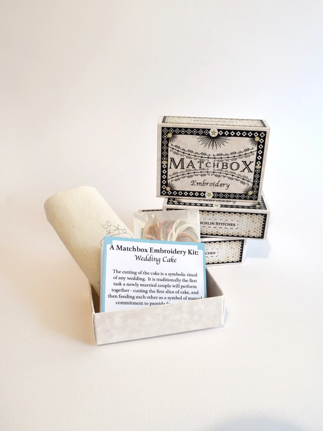 The kit supplied in a beautifully designed matchbox, pre-printed fabric and small instruction leaflet neatly folded to sit inside, along with the yarns and needle housed in a small glassine packet.  A stack of matchboxes sit in the background.