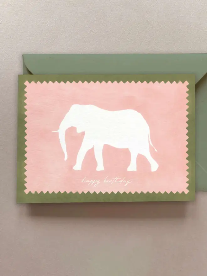 Birthday card featuring a white elephant silhouette on a soft pink background with a zigzag edge detail, bordered in green and paired with a matching green envelope.