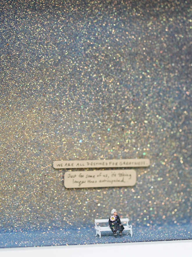 Miniature scene in an artbox showing a tiny old lady sitting on a bench, set against a sparkling purple-blue-grey backdrop (close up). 