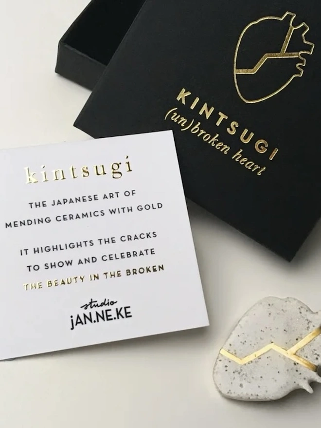(Un)Broken Heart Pin, a white and gold heart pin sitting on a white surface next to its black box and card. 