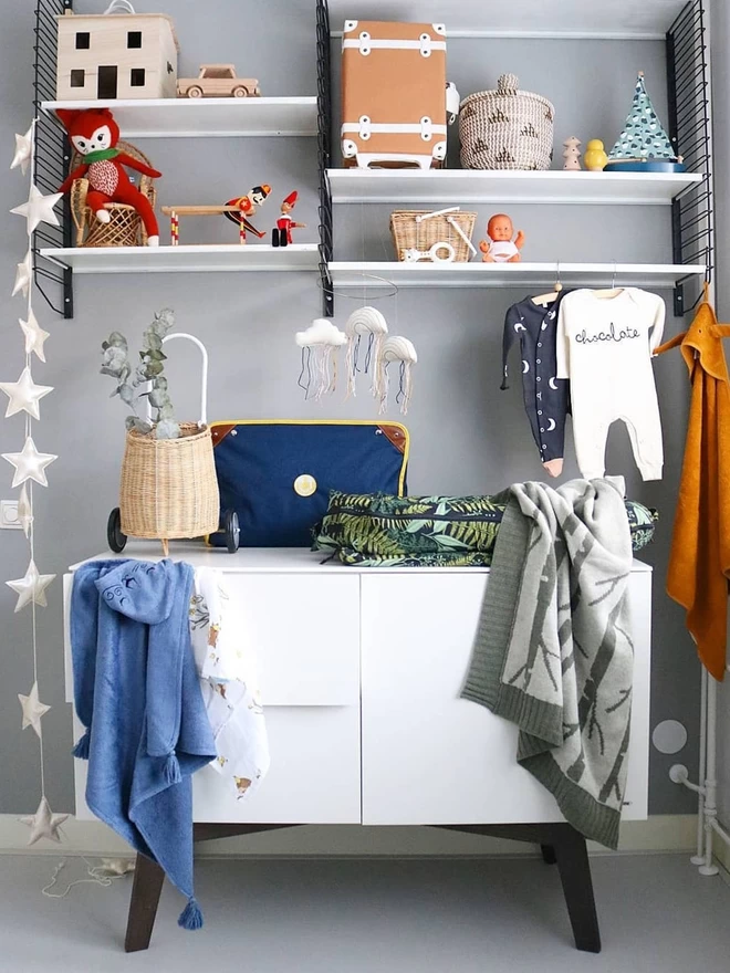 Picture centres on a white chest of drawers with shelves above. The birch forest baby blanket is draped over a changing basket that site on top of the drawers. Toys sit on the shelves and babygrows hang above the balnket.