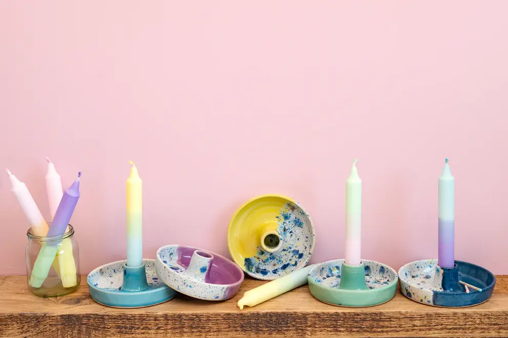 Shoreline Candle Stick Holder, a collection of colourful candle stick holders placed on a wooden shelf. They sit against a pink backdrop. 