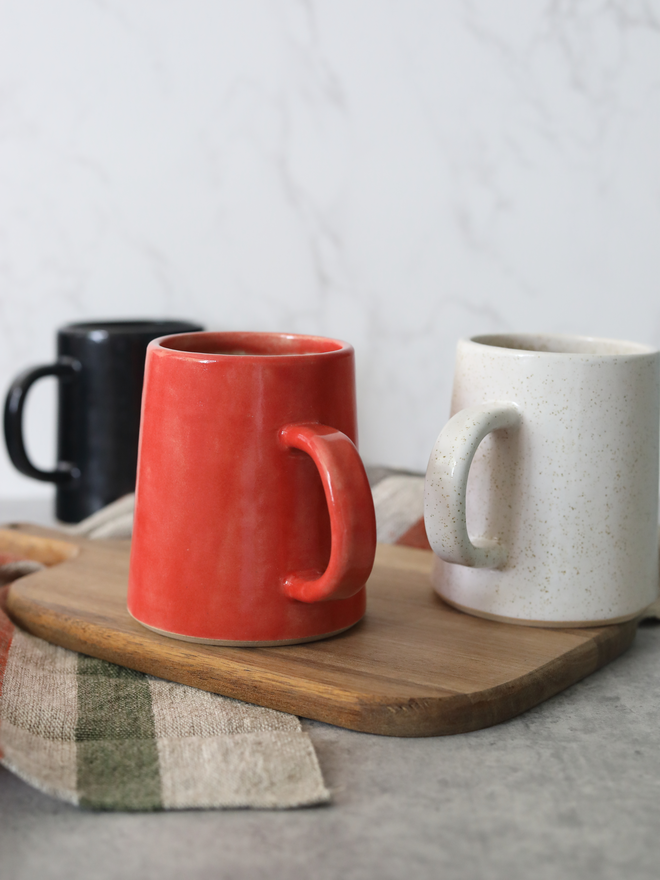 Three handmade ceramic pint mugs on a wooden board — cherry red and speckled white in the foreground, black behind — styled with a green checked linen cloth against a white marble background