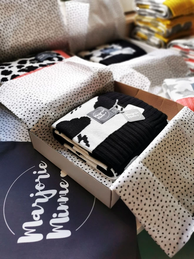 A black and white blanket, neatly folded in a box with tissue paper with a branded gift bag beneath.