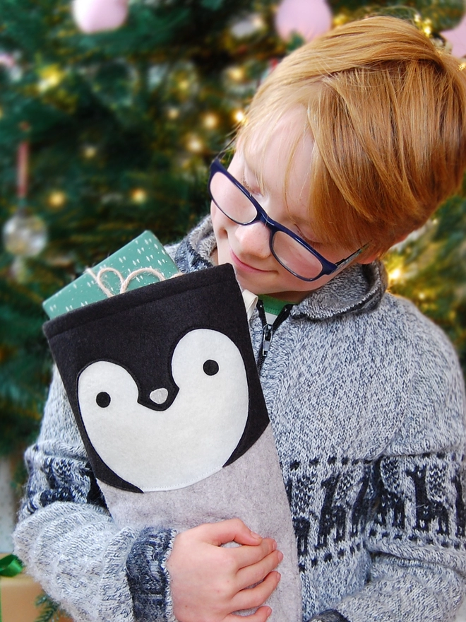 A young boy sitting in front of a Christmas Tree hugs a handmade felt penguin stocking.