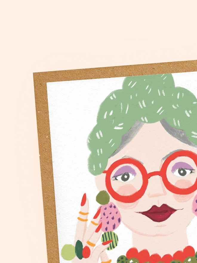 A close up of the texture of a recycled greeting card, illustrated with a fun, glamorous woman in red glasses, red lipstick, a hair scarf and lots of jewellery.