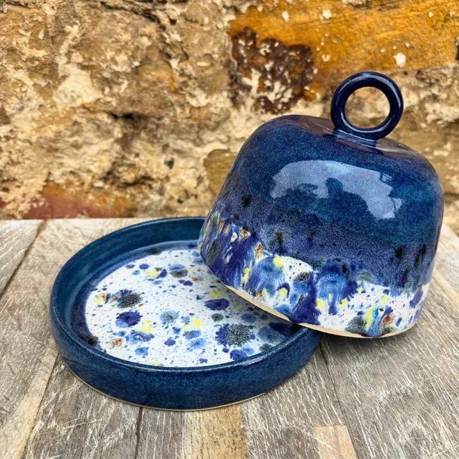 Shoreline Butter Dish, a colourful butter dish placed on a wooden surface against a brick wall. The lid is coming off in the image. 