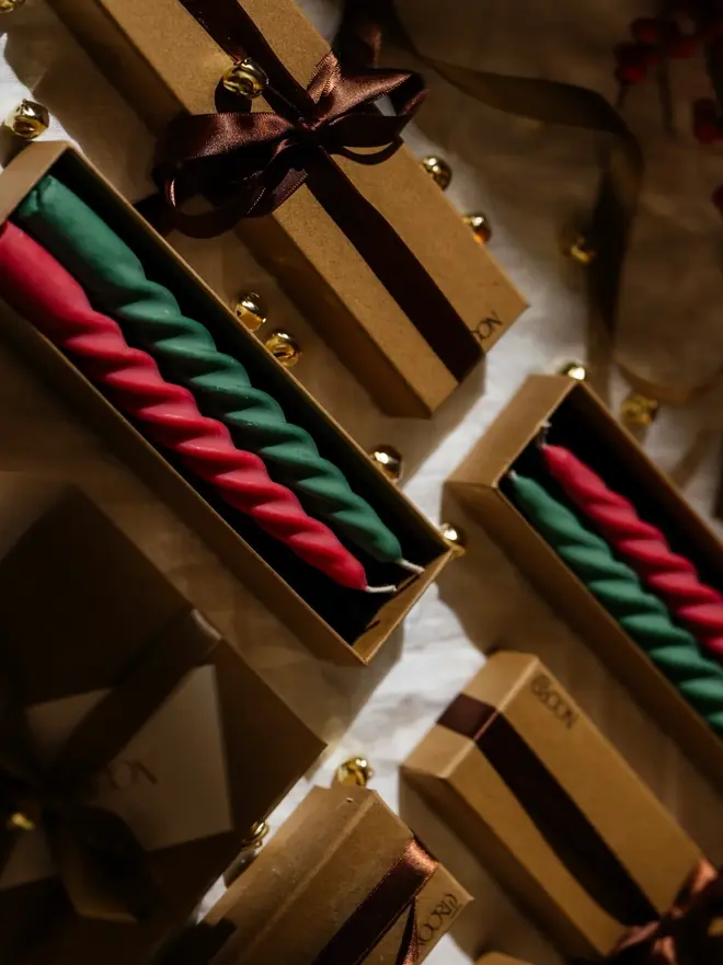 Christmas Spiral Candles Gift Box (Set Of 2). Pairs of red and green twisted taper candles in open kraft Noord gift boxes, surrounded by boxes tied with brown satin ribbon, gold jingle bells and a linen cloth. The gift-ready spiral candles make a festive present or a colourful finishing touch for Christmas tables.