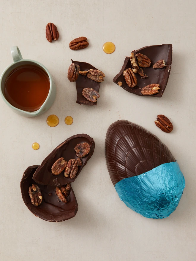 Maple & Pecan Dark Chocolate Easter Egg. a dark chocolate easter egg partially unwrapped from blue foil, photographed broken into pieces with pecan nuts and maple syrup.