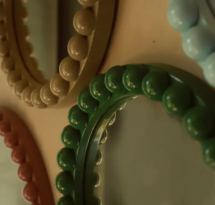 Many round oversized bobbin mirrors, in different colours and sizes with a decorative bobbin-style wooden frame, hanging on a white wall.