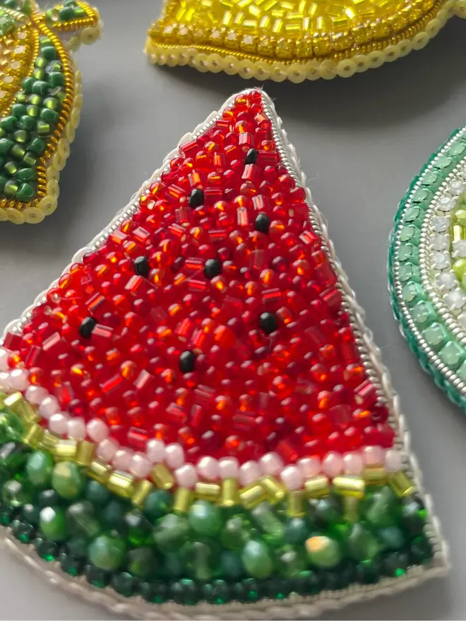Watermelon Beaded Brooch displayed a grey table amongst other beaded fruit brooches including lemons. 