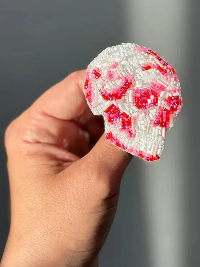 Beaded Pink Skull Brooch  being held in a hand, against a grey background. 