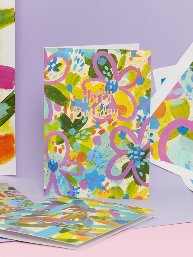 The colourful Raspberry Blossom birthday card sits in front of a lilac backdrop and the paintings used to create its vibrant design