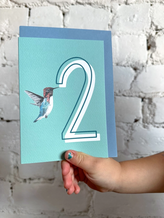 a pale teal card with a big white number 2 and an illustration of a hummingbird with a blue envelope